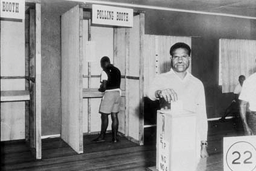 Black and white photo of a man smiling as he casts a vote into a ballot box, while another person fills out a ballot inside a polling booth. The setting is a polling station, conveying a sense of civic duty and participation.