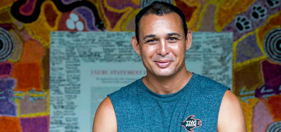 A man in a sleeveless shirt stands smiling in front of a vibrant, patterned Aboriginal artwork. The colorful background adds a warm and cultural tone.