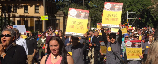A diverse group of people march outdoors, holding colorful signs reading "Our Kids, Our Future" and "Our Culture in Our Hands." The mood is determined.