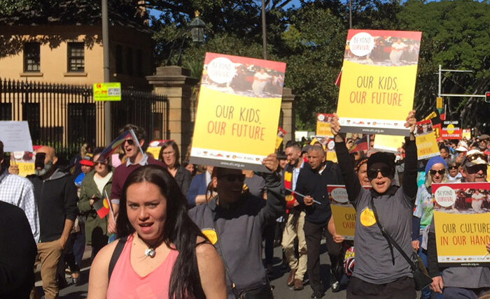 A diverse group of people march outdoors, holding colorful signs reading "Our Kids, Our Future" and "Our Culture in Our Hands." The mood is determined.