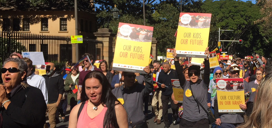 A diverse group of people march outdoors, holding colorful signs reading "Our Kids, Our Future" and "Our Culture in Our Hands." The mood is determined.