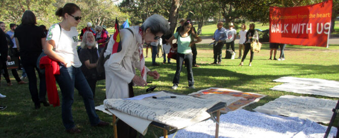A diverse group gathers in a park for a community event. An older woman signs a banner on a table, while others read a "Walk with Us" sign nearby.