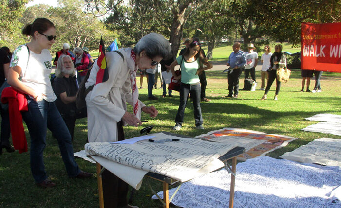 A diverse group gathers in a park for a community event. An older woman signs a banner on a table, while others read a "Walk with Us" sign nearby.
