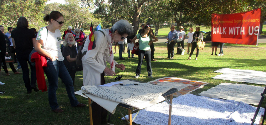 A diverse group gathers in a park for a community event. An older woman signs a banner on a table, while others read a "Walk with Us" sign nearby.