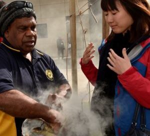 A man performs a traditional smoking ceremony, creating swirling smoke, while a woman observes, engaged and curious. The scene is outdoors with cultural significance.