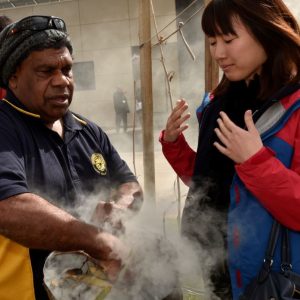 A man performs a traditional smoking ceremony, creating swirling smoke, while a woman observes, engaged and curious. The scene is outdoors with cultural significance.