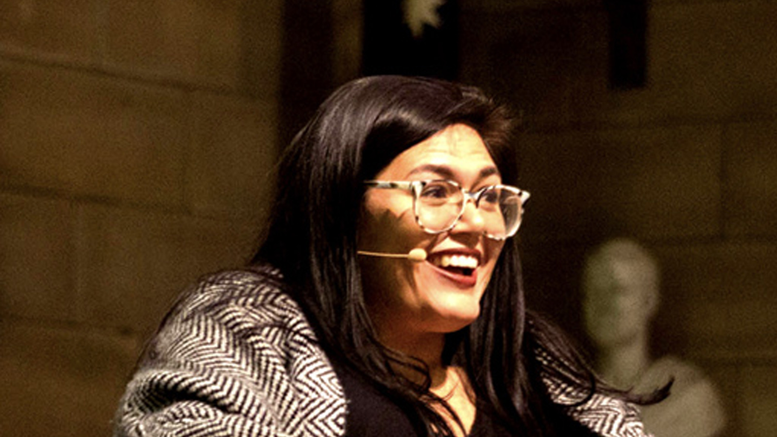A woman with long dark hair and glasses, wearing a microphone and a patterned jacket, smiles widely while speaking in a warmly lit indoor setting.