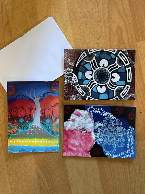 Three colorful postcards with artistic designs—a mandala, a stylized tree scene, and a close-up of a pink bird—are arranged on a wooden surface next to a white envelope.