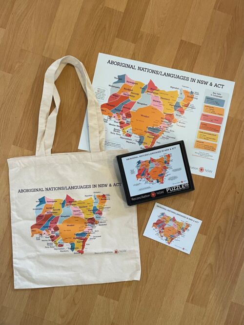A tote bag, puzzle box, poster, and postcard featuring a colorful map of Aboriginal nations and languages in NSW and ACT, laid on a wooden floor.