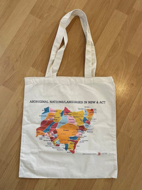 A white tote bag displays a colorful map of Aboriginal nations and languages in NSW and ACT. The text "Reconciliation NSW" is at the bottom.