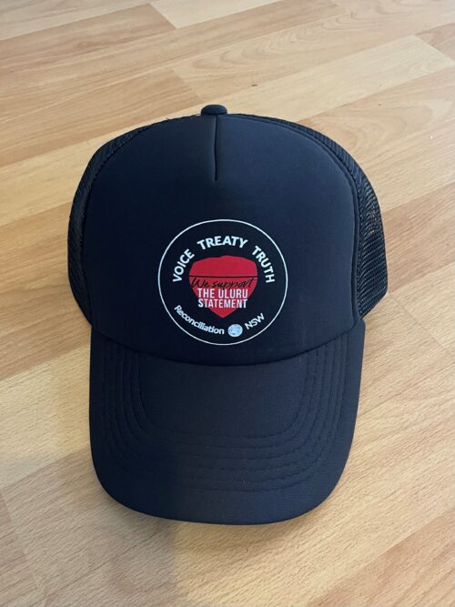 Black trucker cap on a wooden floor, featuring a circular patch with "Voice Treaty Truth" around the edges and "We support the Uluru Statement" in the center.