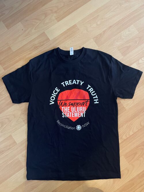 Black T-shirt with "Voice Treaty Truth" in white around a red circle. Text reads "We support the Uluru Statement." Lying on a wood floor.