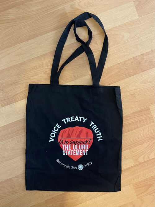 A black Uluru Supporter Tote Bag on a wooden surface. It features a design with the words "Voice Treaty Truth" encircling a red shape, reading "We support the Uluru Statement."