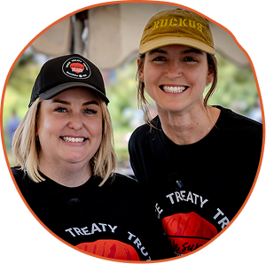 Two people wearing black shirts and caps smile warmly under a canopy. Their shirts and hats have logos and text. The scene is bright and cheerful.